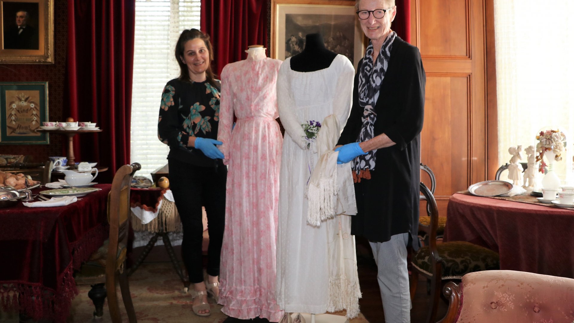 ​Caption: Heritage Facilities Curator Māhina Marshall and Textile Technician volunteer Karen Richards with two of the garments featuring in the Secret Lives of Dresses.